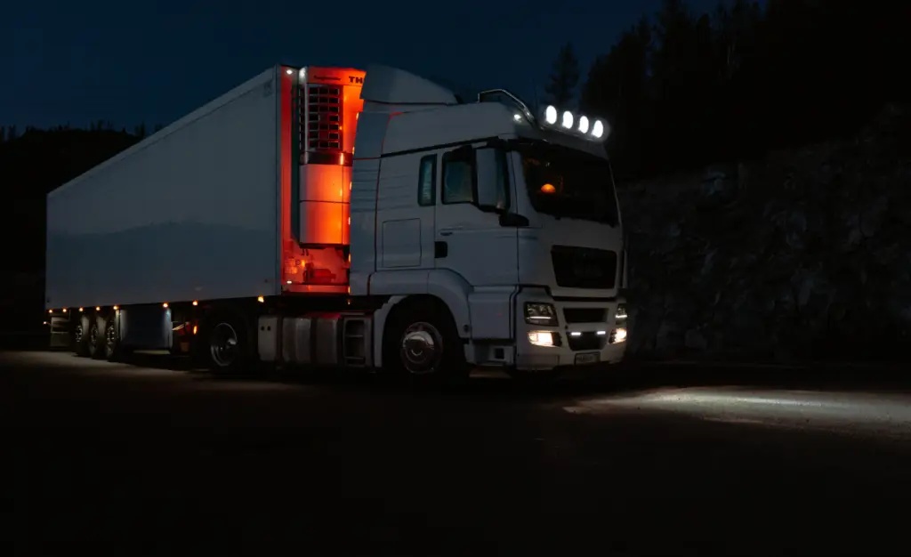 Rapid Truck Repair - White semi-truck with trailer parked on a dark road, headlights and roof lights on, orange interior trailer lights glowing.
