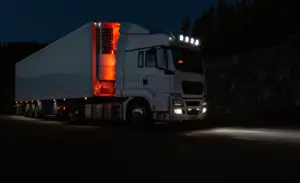Rapid Truck Repair - White semi-truck with trailer parked on a dark road, headlights and roof lights on, orange interior trailer lights glowing.
