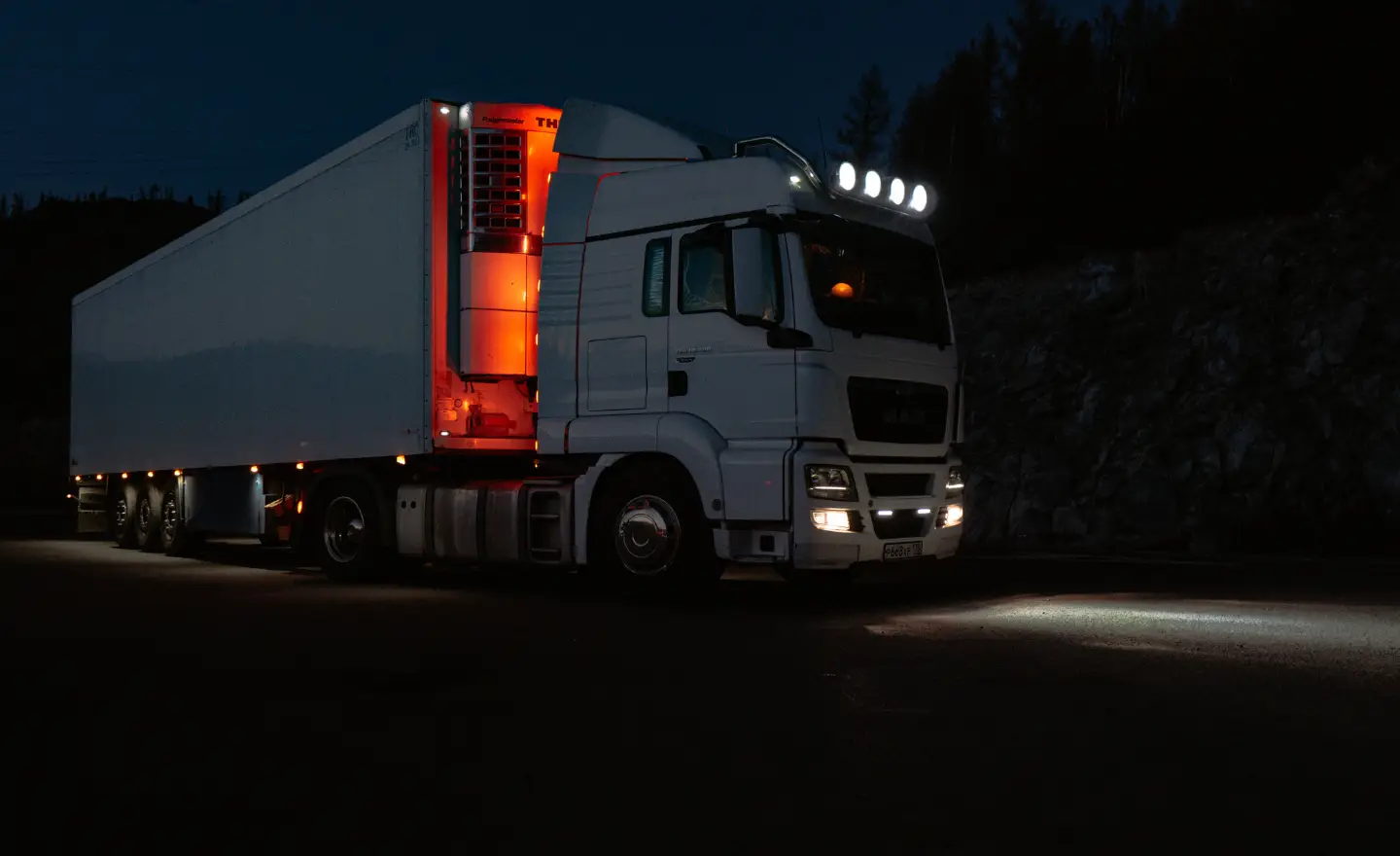 247 Truck Roadside Assistance Joplin MO Mobile Help at the Crossroads of I-44 I-49 - Rapid Truck Repair - Rapid Truck Repair Rapid Truck Repair - Rapid Truck Repair - White semi-truck with trailer parked on a dark road, headlights and roof lights on, orange interior trailer lights glowing.