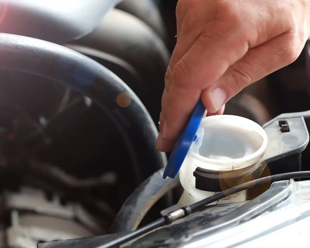 Rapid Truck Repair - Hand lifting a blue cap off a white coolant reservoir in a car engine bay.